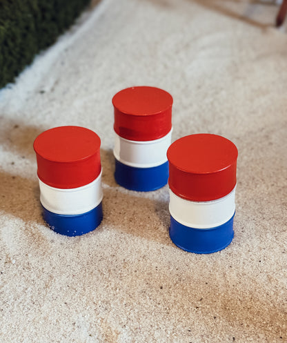 Three small cylindrical stacked items painted red, white, and blue, placed on a light sandy surface.