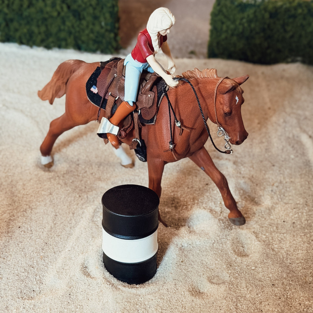 A miniature toy figurine of a woman riding a brown horse, positioned in a sandy arena with a black and white barrel in the foreground.