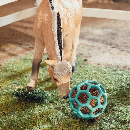 A small calf stands in a barn-like enclosure on artificial grass, eating hay while a teal ball-shaped hay toy lies beside it. A white fence and out-of-focus barn interior are visible in the background.