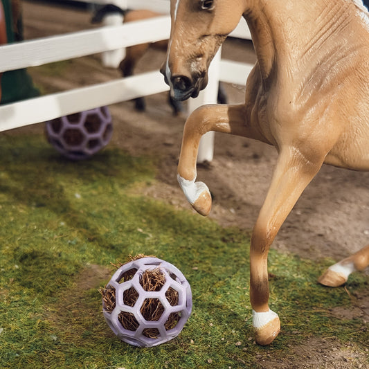 A tan horse toy or model horse standing beside a purple honeycomb ball filled with dried grass on artificial turf, with a white fence and another purple ball in the background.