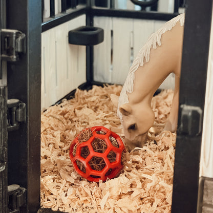 A beige horse figurine with white decorative details is shown inside a miniature stable, sniffing a red ball toy on a bed of wood shavings.