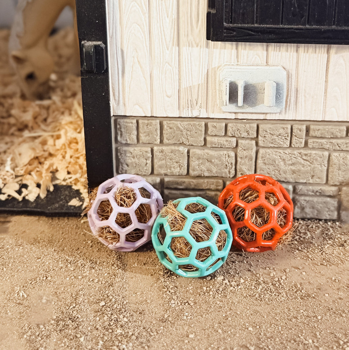 Three small plastic ball toys for small animals, featuring purple, mint green, and orange colors, filled with natural straw. They are placed on sandy ground in front of a miniature white wooden barn.