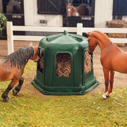 Two Schleich model horses feeding from opposite sides of the green hay bell feeder in a stable diorama