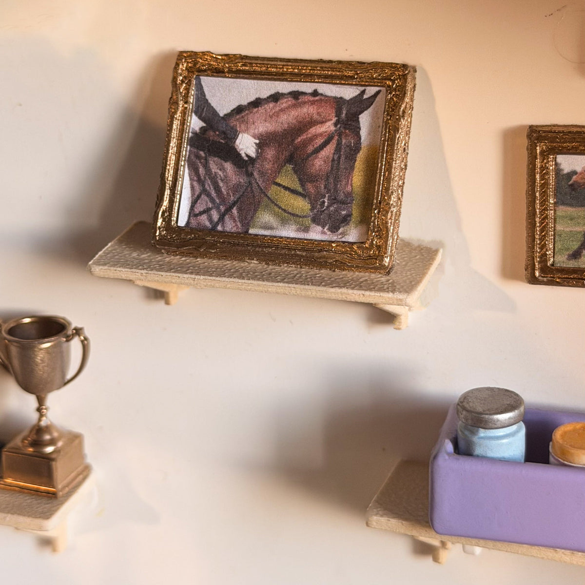 Gold miniature frame with a dressage horse picture standing on a small wooden shelf, with a trophy and a lavender box on the shelves below