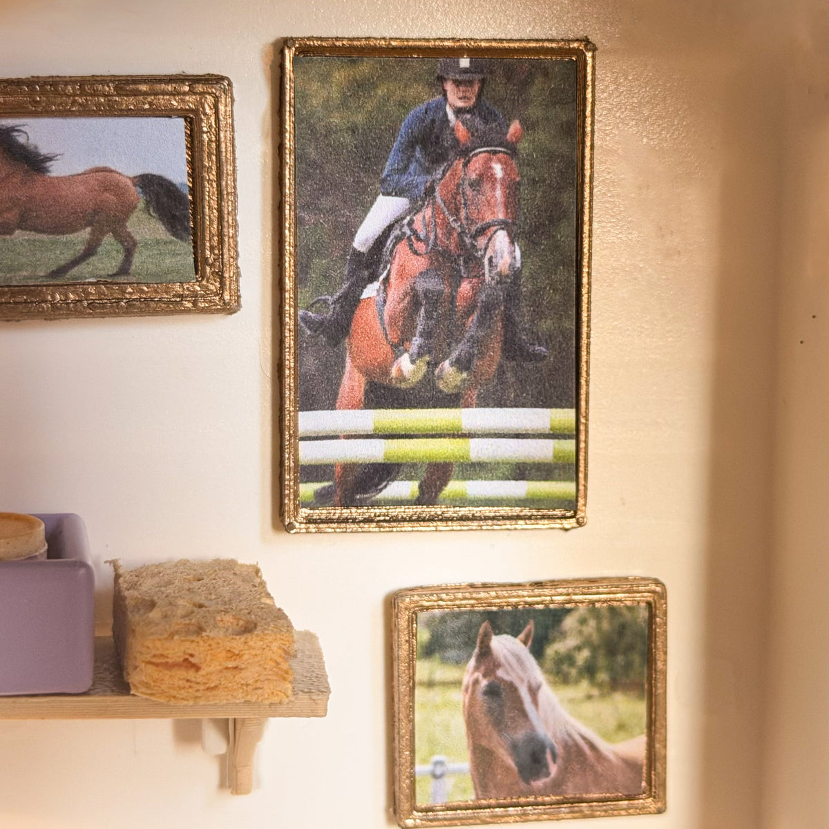 Gold miniature frames with a show-jumping horse picture and a chestnut horse portrait hanging on a cream wall above a small shelf