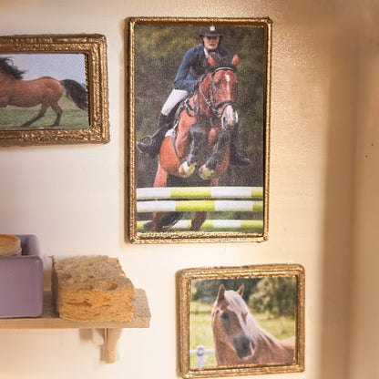 Gold miniature frames with a show-jumping horse picture and a chestnut horse portrait hanging on a cream wall above a small shelf