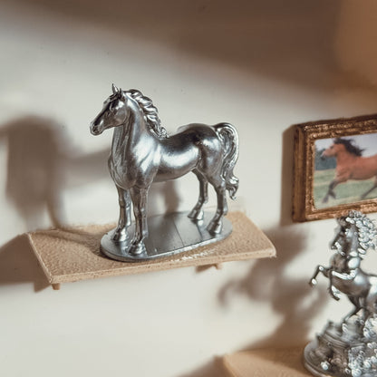 Silver standing horse statue on an oval base on a miniature shelf, with a framed horse picture and the rearing horse statue in the background