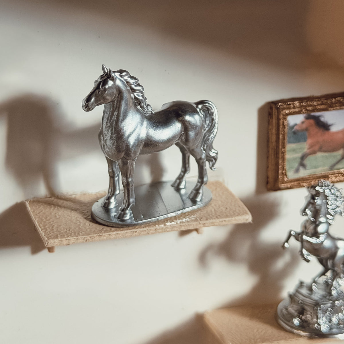 Silver standing horse statue on an oval base displayed on a miniature wall shelf next to a framed horse picture