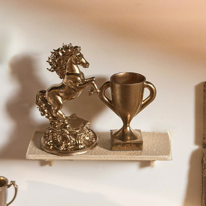 Gold rearing horse statue on an ornate base next to a gold trophy cup on a miniature shelf