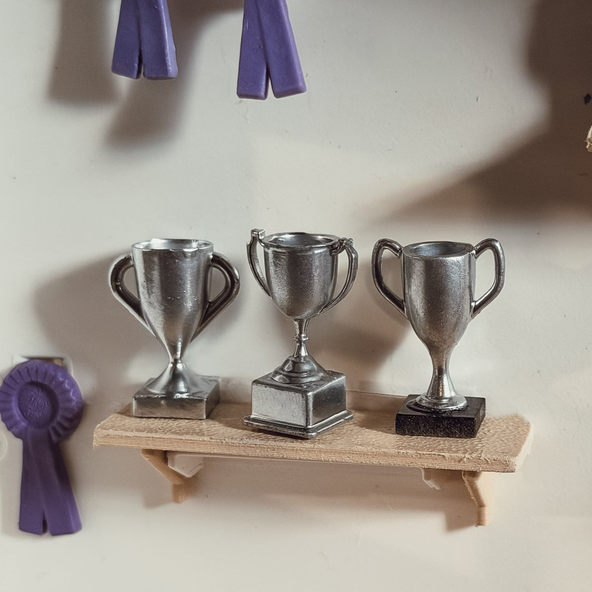 Three silver miniature trophy cups with different handles and bases on a small wooden shelf, next to purple rosettes