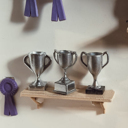 Three silver miniature trophy cups with different handles and bases on a small wooden shelf, next to purple rosettes