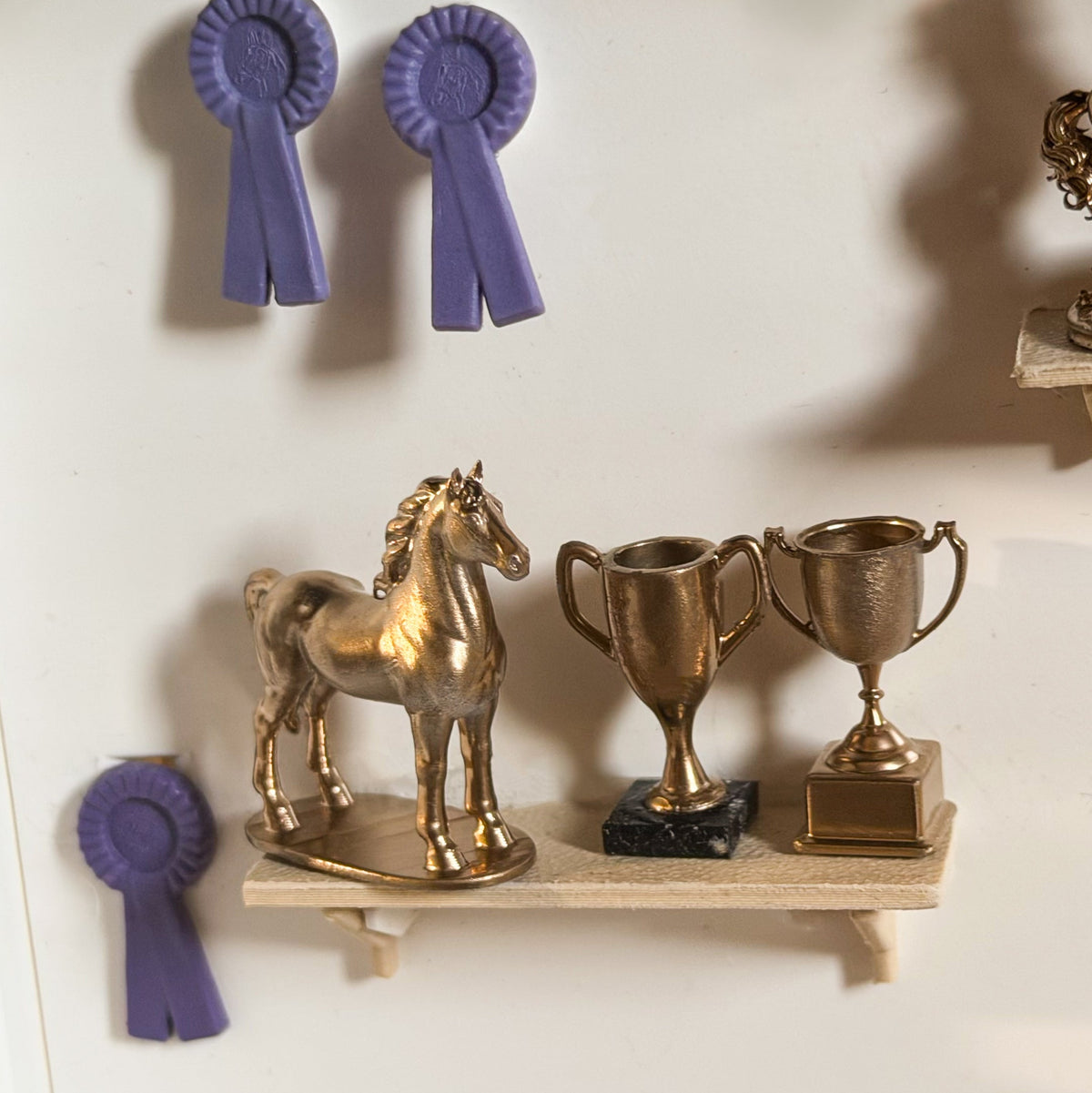 Gold standing horse statue and two gold trophy cups on a miniature wooden shelf, with purple rosettes on the wall