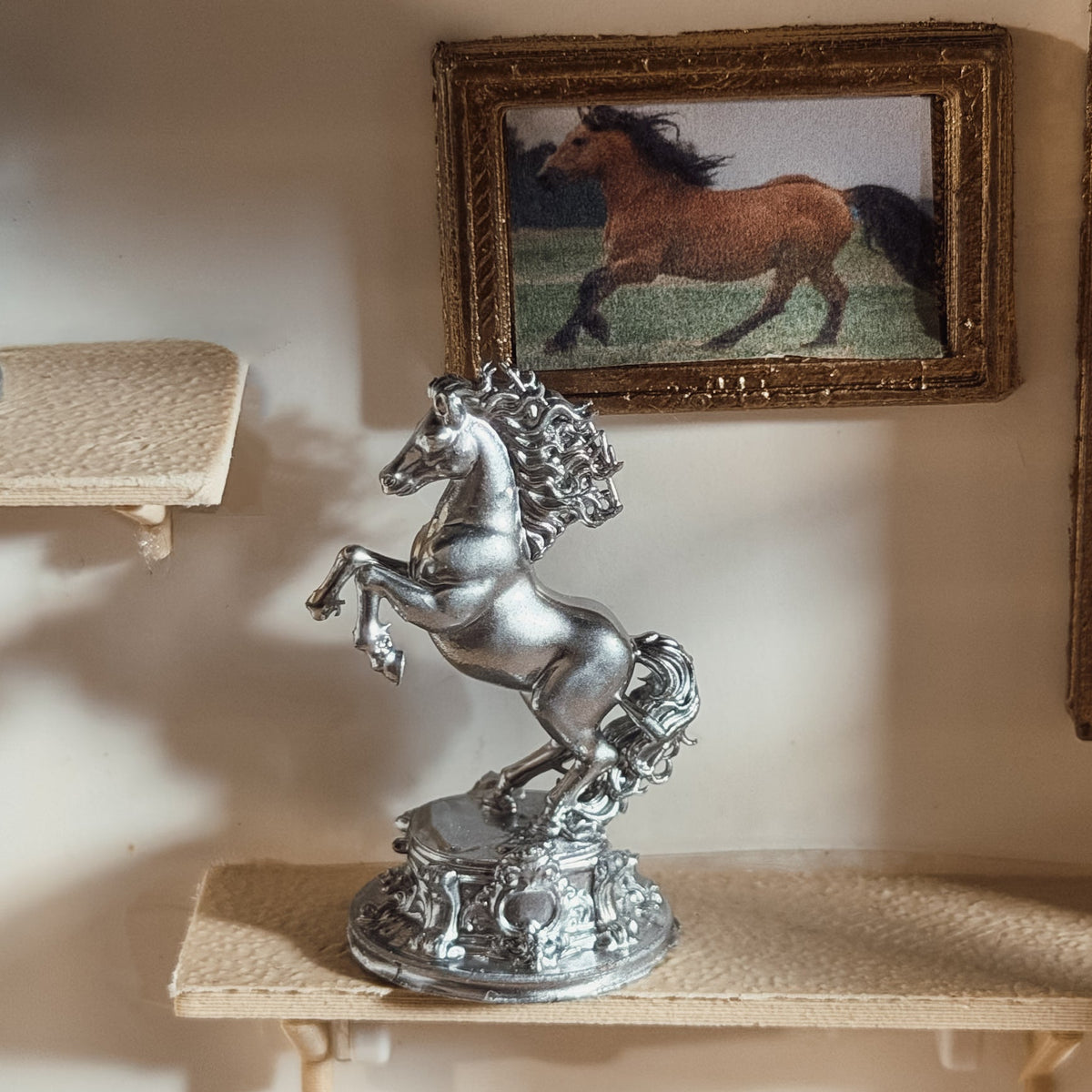 Silver rearing horse statue on an ornate base displayed on a miniature shelf below a framed horse picture